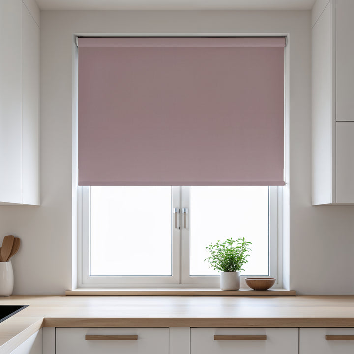 Pink roller blind on a window in a kitchen with white cabinets and a plant on the windowsill.