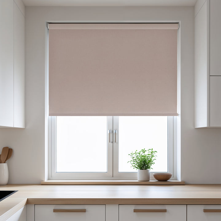 Beige roller blind on a window in a kitchen with white cabinets and a plant on the windowsill.